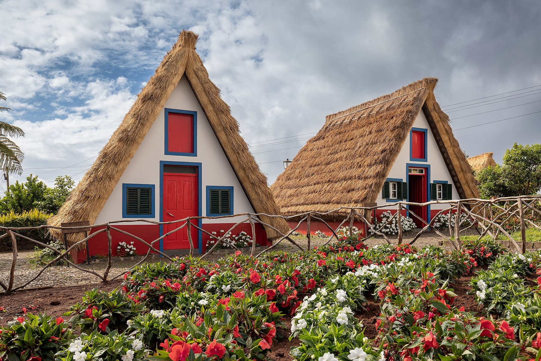 Two traditional triangular houses in Santana with steep thatched roofs, red doors, blue trim and flower beds.