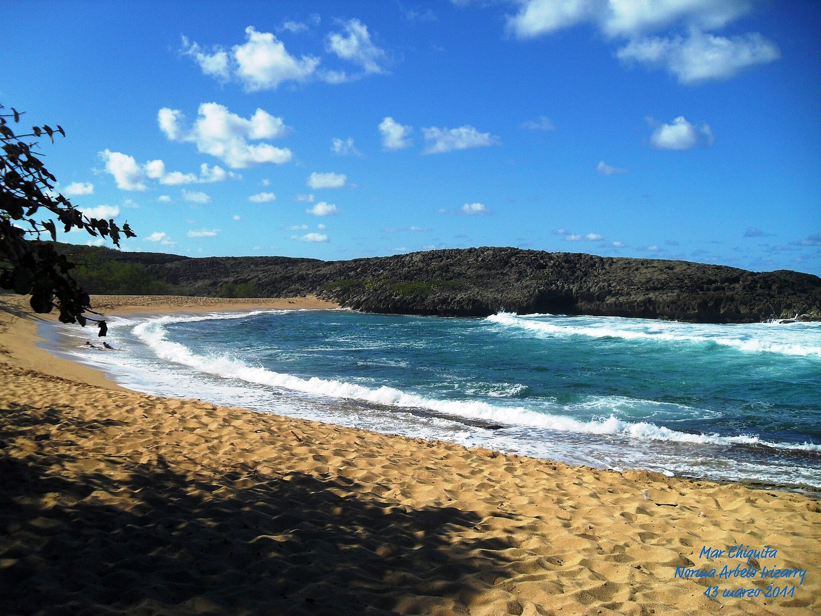 Sunlit golden sand and blue water inside Mar Chiquita’s enclosed bay, with a limestone ridge around the cove.