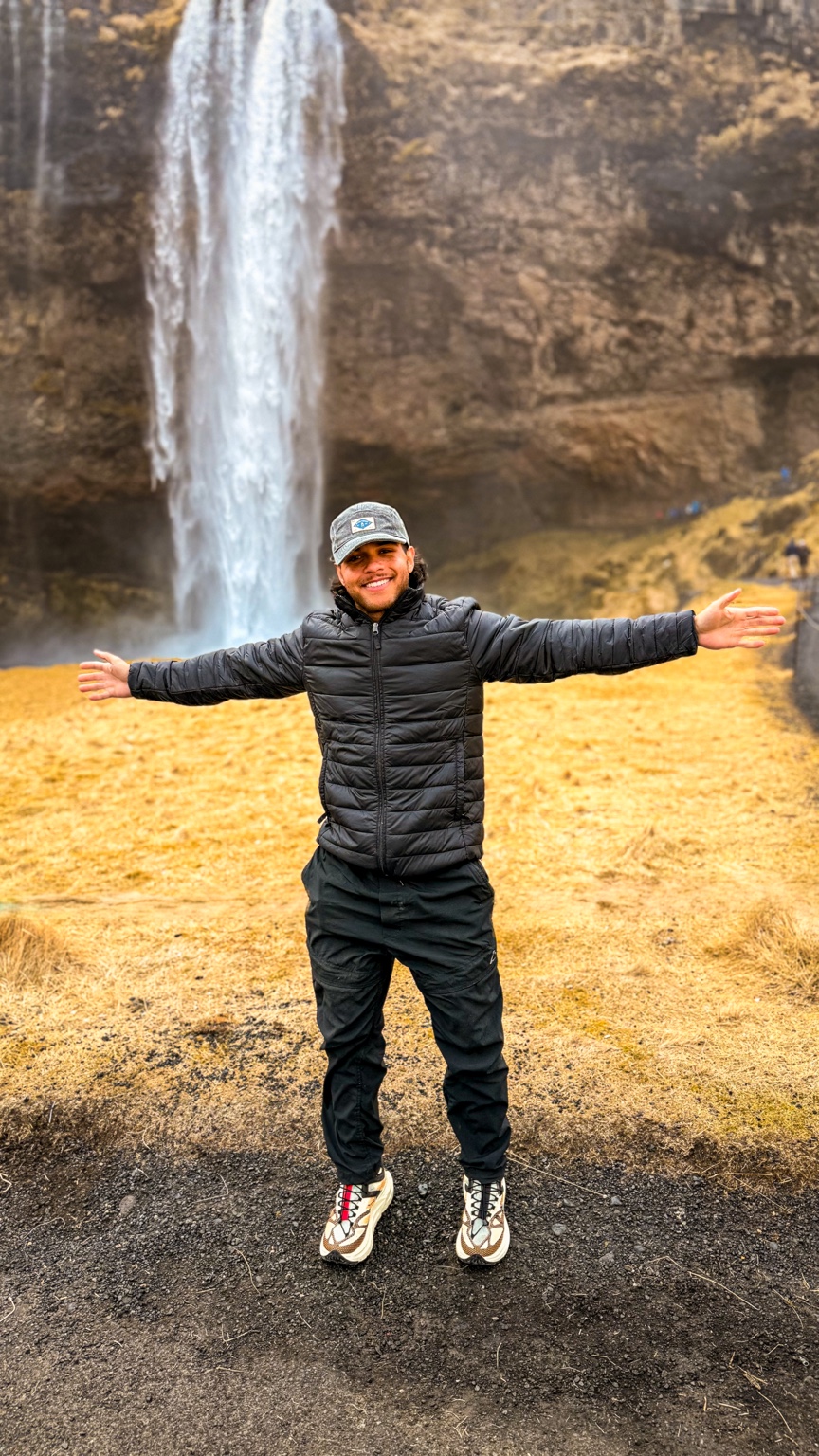 Sebastian smiling with his arms outstretched in front of a waterfall